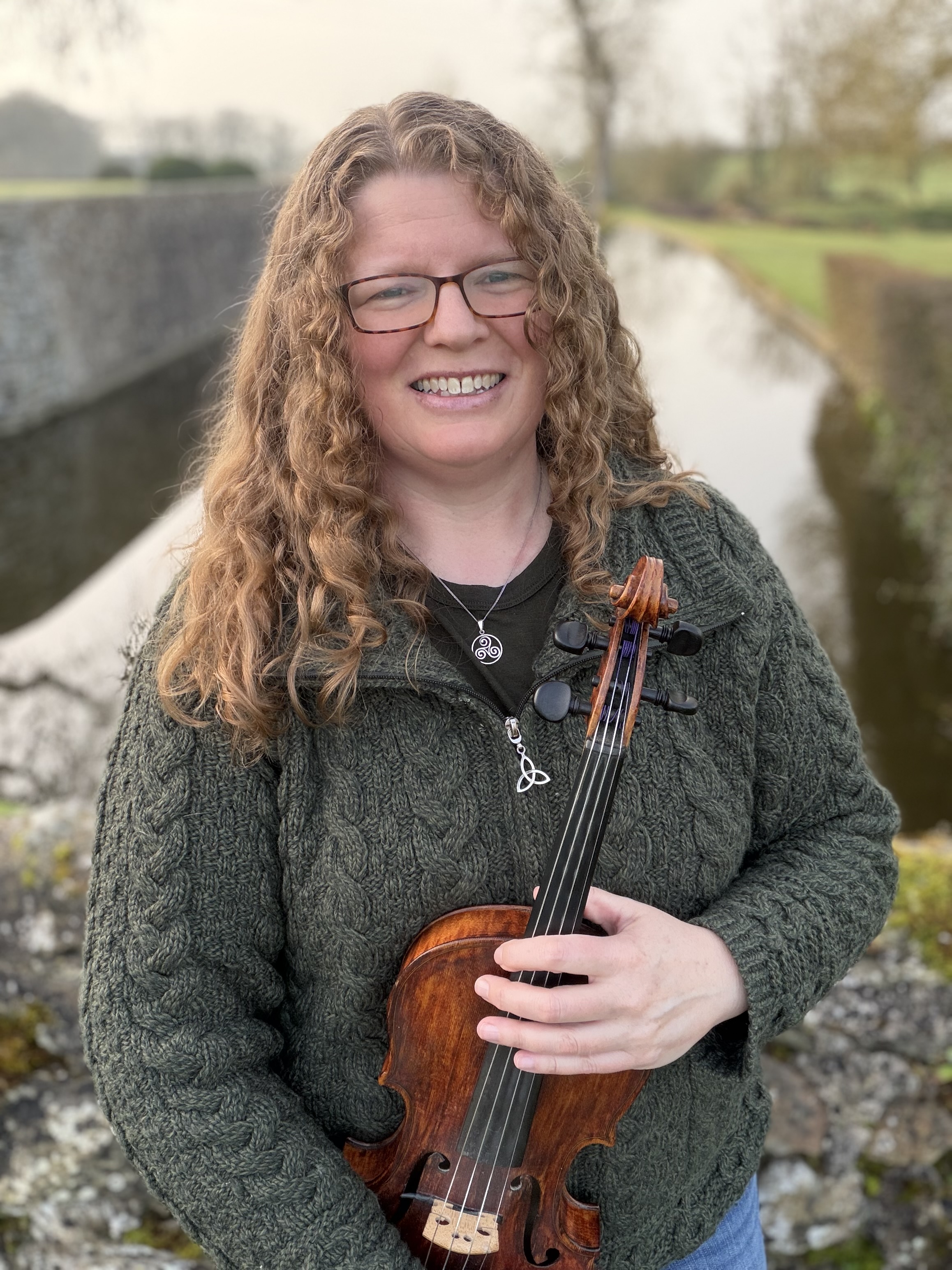 Katherine Moller standing in front of river holding her violin while smiling
